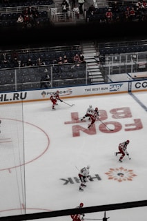 A hockey game scene featuring players in red and black jerseys skating on an ice rink. The rink is set inside an arena with partially occupied spectator stands. Visible advertisements along the rink boards indicate sponsorships.