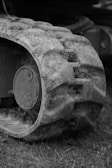 A close-up of a rugged, muddy track of a vehicle or machinery with visible dirt and grass. The texture details on the track exhibit signs of wear and use, suggesting it has been operated in an outdoor environment. The background is slightly blurred, emphasizing the focus on the track.