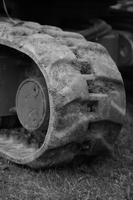 A close-up of a rugged, muddy track of a vehicle or machinery with visible dirt and grass. The texture details on the track exhibit signs of wear and use, suggesting it has been operated in an outdoor environment. The background is slightly blurred, emphasizing the focus on the track.