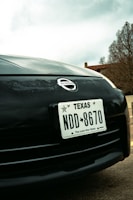 Close-up view of a car's front end featuring a Texas license plate with the number NDD 8670. The Nissan logo is prominently displayed above the license plate. In the background, there are trees and part of a building, with an overcast sky.