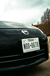 Close-up view of a car's front end featuring a Texas license plate with the number NDD 8670. The Nissan logo is prominently displayed above the license plate. In the background, there are trees and part of a building, with an overcast sky.