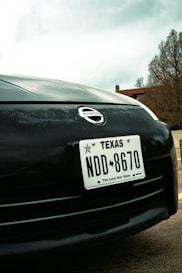 Close-up view of a car's front end featuring a Texas license plate with the number NDD 8670. The Nissan logo is prominently displayed above the license plate. In the background, there are trees and part of a building, with an overcast sky.