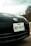 Close-up view of a car's front end featuring a Texas license plate with the number NDD 8670. The Nissan logo is prominently displayed above the license plate. In the background, there are trees and part of a building, with an overcast sky.
