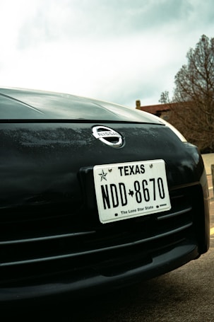 Close-up view of a car's front end featuring a Texas license plate with the number NDD 8670. The Nissan logo is prominently displayed above the license plate. In the background, there are trees and part of a building, with an overcast sky.