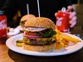A cheeseburger with a sesame seed bun is placed on a white plate, accompanied by a side of fries. The burger is stacked with layers including a beef patty with melted cheese, lettuce, tomato, onion, and a fried egg on top. Pickles are visible on the side of the plate. There is a can of soda and another burger in the background.
