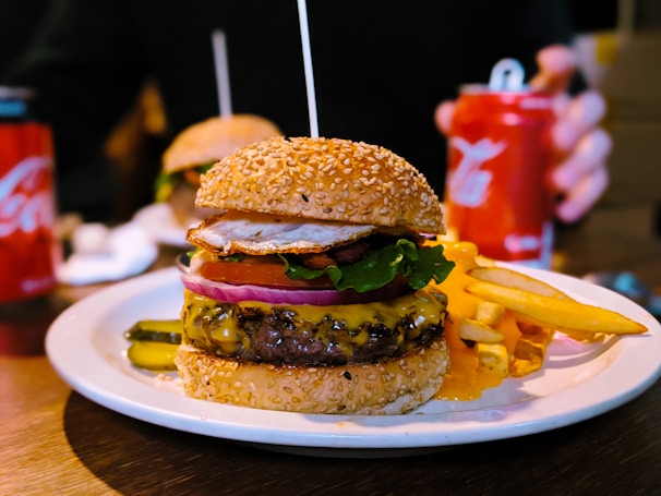A cheeseburger with a sesame seed bun is placed on a white plate, accompanied by a side of fries. The burger is stacked with layers including a beef patty with melted cheese, lettuce, tomato, onion, and a fried egg on top. Pickles are visible on the side of the plate. There is a can of soda and another burger in the background.