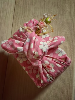 A fabric-wrapped object with a pink floral pattern is adorned with small artificial flowers and tied with a decorative knot. The background features a light wooden surface, adding a natural contrast to the colorful fabric.