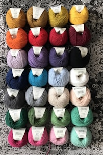 Several rows of yarn balls with labels are arranged on a patterned fabric background. The yarn comes in a variety of colors including yellow, red, blue, purple, green, and pink. Each ball has a label on top indicating its brand and type.