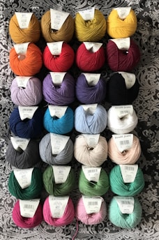 Several rows of yarn balls with labels are arranged on a patterned fabric background. The yarn comes in a variety of colors including yellow, red, blue, purple, green, and pink. Each ball has a label on top indicating its brand and type.