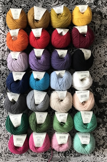 Several rows of yarn balls with labels are arranged on a patterned fabric background. The yarn comes in a variety of colors including yellow, red, blue, purple, green, and pink. Each ball has a label on top indicating its brand and type.