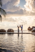 A family stands on a boardwalk by the water, silhouetted against a dramatic sky with large, fluffy clouds. Palm fronds are visible on the left, and overwater bungalows are seen in the background, evoking a tropical paradise.