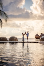A family stands on a boardwalk by the water, silhouetted against a dramatic sky with large, fluffy clouds. Palm fronds are visible on the left, and overwater bungalows are seen in the background, evoking a tropical paradise.