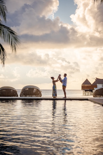 A family stands on a boardwalk by the water, silhouetted against a dramatic sky with large, fluffy clouds. Palm fronds are visible on the left, and overwater bungalows are seen in the background, evoking a tropical paradise.