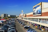 A bustling shopping area with a large building featuring billboards is shown. The scene includes a busy parking lot filled with numerous cars and people walking along the sidewalk, browsing market stalls set up under the building's overhang. Mid-rise apartment buildings and trees are visible in the background under a clear blue sky.