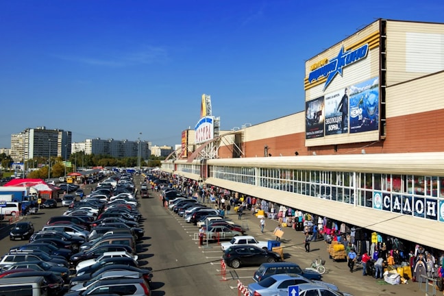 A bustling shopping area with a large building featuring billboards is shown. The scene includes a busy parking lot filled with numerous cars and people walking along the sidewalk, browsing market stalls set up under the building's overhang. Mid-rise apartment buildings and trees are visible in the background under a clear blue sky.