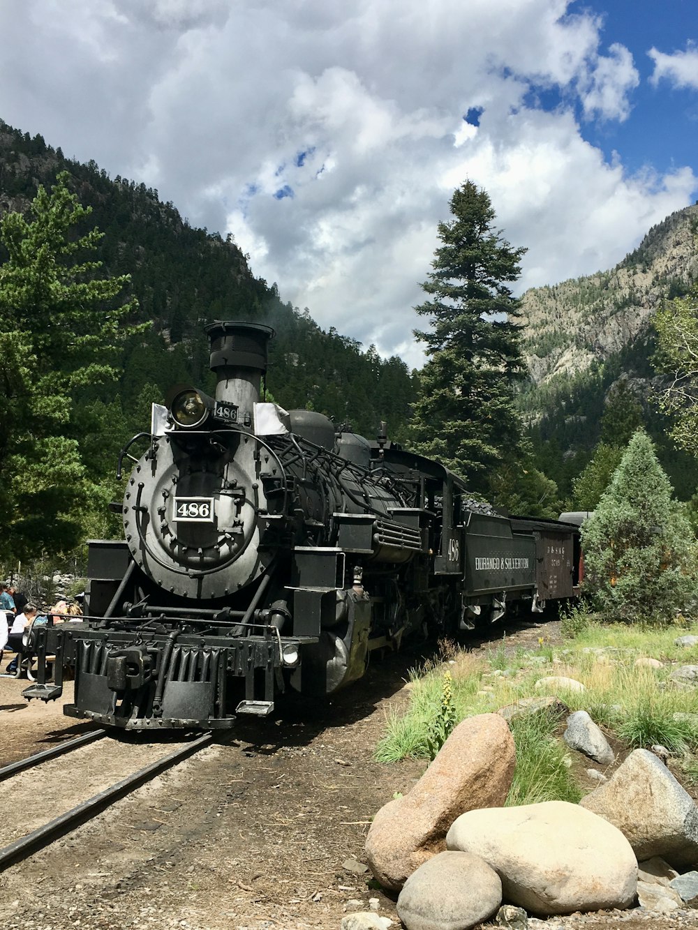 black train on rail tracks near green trees under white clouds and blue sky during daytime