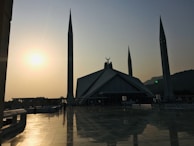 A majestic mosque with multiple tall minarets is seen at sunset, casting a warm glow over the reflective tiles of a spacious courtyard. The sun is low in the sky, creating a silhouette effect on the structure. Trees and a mountain are visible in the background, enhancing the serene ambiance.