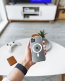 A hand holds a smartphone with a case featuring a prominent circular design and Apple logo. In the background, there is a modern living room with a white entertainment unit, a large TV screen, and a gaming controller on a white coffee table.