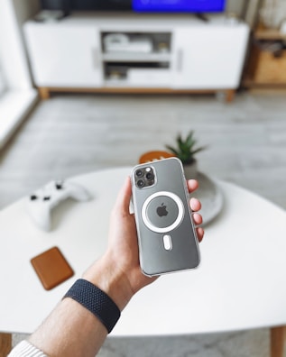 A hand holds a smartphone with a case featuring a prominent circular design and Apple logo. In the background, there is a modern living room with a white entertainment unit, a large TV screen, and a gaming controller on a white coffee table.