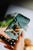 A person holds a smartphone displaying an image of cookies and a whisk on a white surface. The phone's screen is focused on a baking scene, and the background is blurred, showing greenery.