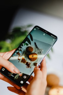 A person holds a smartphone displaying an image of cookies and a whisk on a white surface. The phone's screen is focused on a baking scene, and the background is blurred, showing greenery.