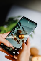A person holds a smartphone displaying an image of cookies and a whisk on a white surface. The phone's screen is focused on a baking scene, and the background is blurred, showing greenery.
