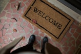A doormat with the word 'WELCOME' is placed on a tiled floor with red and pink geometric tiles. Several dry brown leaves are scattered across the floor. A person wearing black shoes and dark pants is partially visible from an overhead perspective.