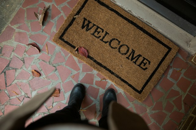 A doormat with the word 'WELCOME' is placed on a tiled floor with red and pink geometric tiles. Several dry brown leaves are scattered across the floor. A person wearing black shoes and dark pants is partially visible from an overhead perspective.