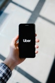 A hand holding a smartphone displaying the Uber logo on its screen. The background appears softly blurred, providing a contrast to the sharp focus on the phone. The hand is wearing a long-sleeved, checkered shirt.
