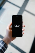 A hand holding a smartphone displaying the Uber logo on its screen. The background appears softly blurred, providing a contrast to the sharp focus on the phone. The hand is wearing a long-sleeved, checkered shirt.