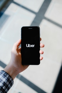A hand holding a smartphone displaying the Uber logo on its screen. The background appears softly blurred, providing a contrast to the sharp focus on the phone. The hand is wearing a long-sleeved, checkered shirt.