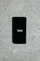 A smartphone displaying the Uber logo is placed on a concrete surface. The phone screen is black with the white Uber logo in the center.