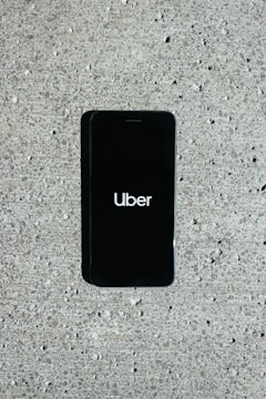 A smartphone displaying the Uber logo is placed on a concrete surface. The phone screen is black with the white Uber logo in the center.