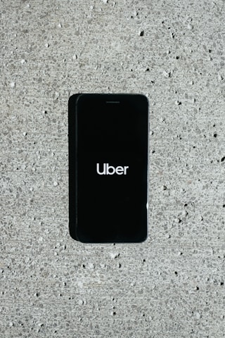 A smartphone displaying the Uber logo is placed on a concrete surface. The phone screen is black with the white Uber logo in the center.