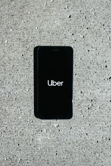 A smartphone displaying the Uber logo is placed on a concrete surface. The phone screen is black with the white Uber logo in the center.