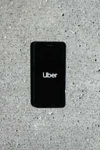 A smartphone displaying the Uber logo is placed on a concrete surface. The phone screen is black with the white Uber logo in the center.