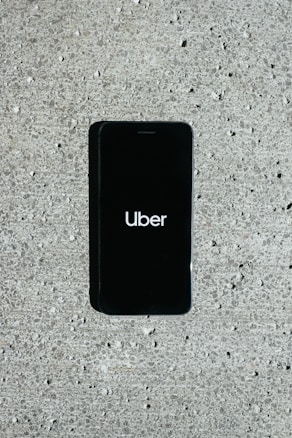 A smartphone displaying the Uber logo is placed on a concrete surface. The phone screen is black with the white Uber logo in the center.
