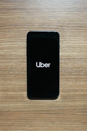 A smartphone with a black screen displays the Uber logo in white text. The phone is placed on a wooden surface with a light brown texture.