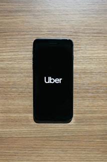 A smartphone with a black screen displays the Uber logo in white text. The phone is placed on a wooden surface with a light brown texture.