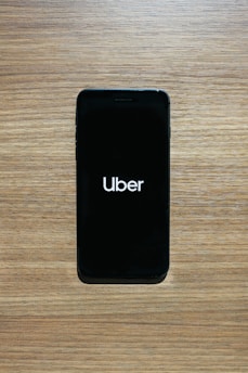 A smartphone with a black screen displays the Uber logo in white text. The phone is placed on a wooden surface with a light brown texture.
