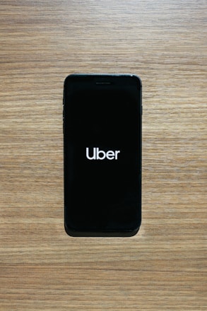 A smartphone with a black screen displays the Uber logo in white text. The phone is placed on a wooden surface with a light brown texture.