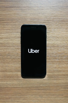 A smartphone with a black screen displays the Uber logo in white text. The phone is placed on a wooden surface with a light brown texture.