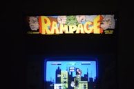An arcade machine featuring the classic game 'Rampage', with its title in bold, colorful letters surrounded by graphics of cartoon-like monsters. The screen displays a scene from the game with pixelated characters and buildings, and the words 'Game Over'.