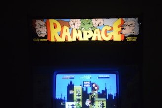 An arcade machine featuring the classic game 'Rampage', with its title in bold, colorful letters surrounded by graphics of cartoon-like monsters. The screen displays a scene from the game with pixelated characters and buildings, and the words 'Game Over'.