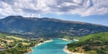 A serene landscape featuring a turquoise river winding through lush green hills, with a backdrop of rolling mountains under a partly cloudy sky. The river creates a beautiful contrast against the vibrant greenery, suggesting a peaceful and untouched natural environment.