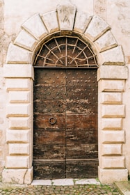 A rustic, arched wooden door with metal studs and a central ring handle, set within a stone wall. The arch features a keystone engraved with the letter 'A' and the number '19'. The surrounding stonework has a textured, weathered appearance, adding to the historic charm.
