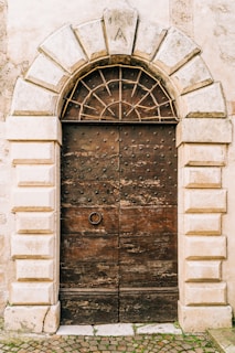 A rustic, arched wooden door with metal studs and a central ring handle, set within a stone wall. The arch features a keystone engraved with the letter 'A' and the number '19'. The surrounding stonework has a textured, weathered appearance, adding to the historic charm.