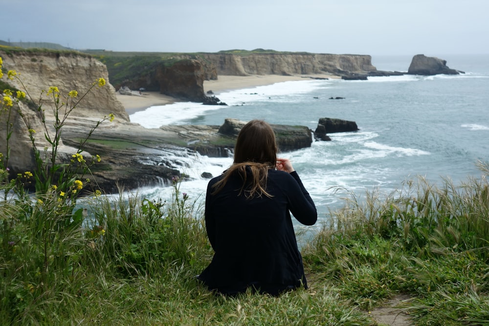 woman in black coat standing on green grass field near body of water during daytime