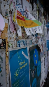 Various torn and layered paper advertisements are posted on a bulletin board. The papers are overlapping, with some of them slightly peeling off. The materials include promotional flyers and posters, featuring different colors and text. One visible blue poster reads 'Ullev&aring;l Barnegospel.'