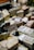 A variety of cheeses are displayed on a market stand, each labeled with pricing. Some of the packages are wrapped in plastic. There are visible price tags, including one for 'Pecorino' and others with varying prices. In the background, there are bottles and other produce items.